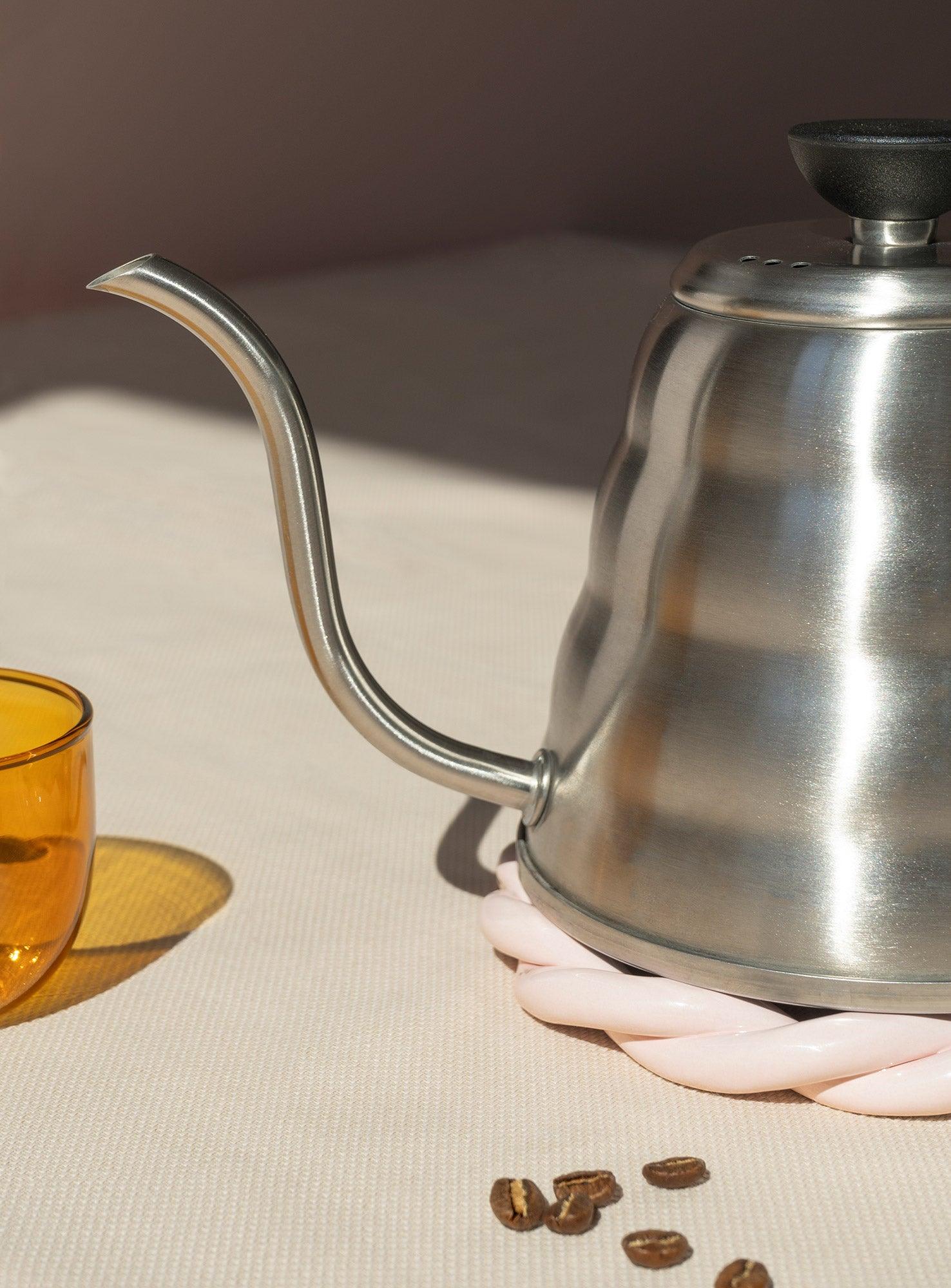 A pink OCTAEVO Sicilia Ceramic Trivet holds a gooseneck kettle near a yellow glass and coffee beans on beige.