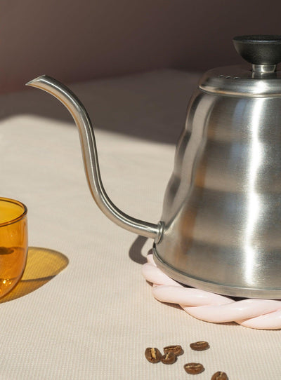 A pink OCTAEVO Sicilia Ceramic Trivet holds a gooseneck kettle near a yellow glass and coffee beans on beige.