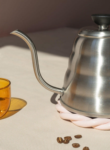 A pink OCTAEVO Sicilia Ceramic Trivet holds a gooseneck kettle near a yellow glass and coffee beans on beige.