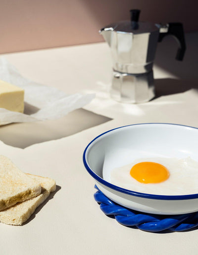 A bowl, toast, egg, butter, and a stovetop espresso maker sit on a blue OCTAEVO Sicilia Ceramic Trivet.