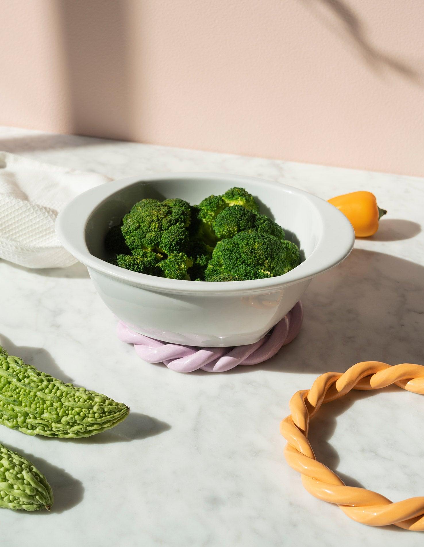 A white bowl of steamed broccoli rests on a marble surface with OCTAEVO purple Sicilia Ceramic Trivet. 