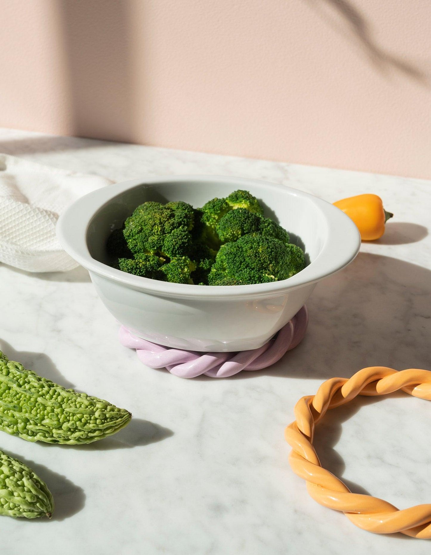 A white bowl of steamed broccoli rests on a marble surface with OCTAEVO purple Sicilia Ceramic Trivet. 