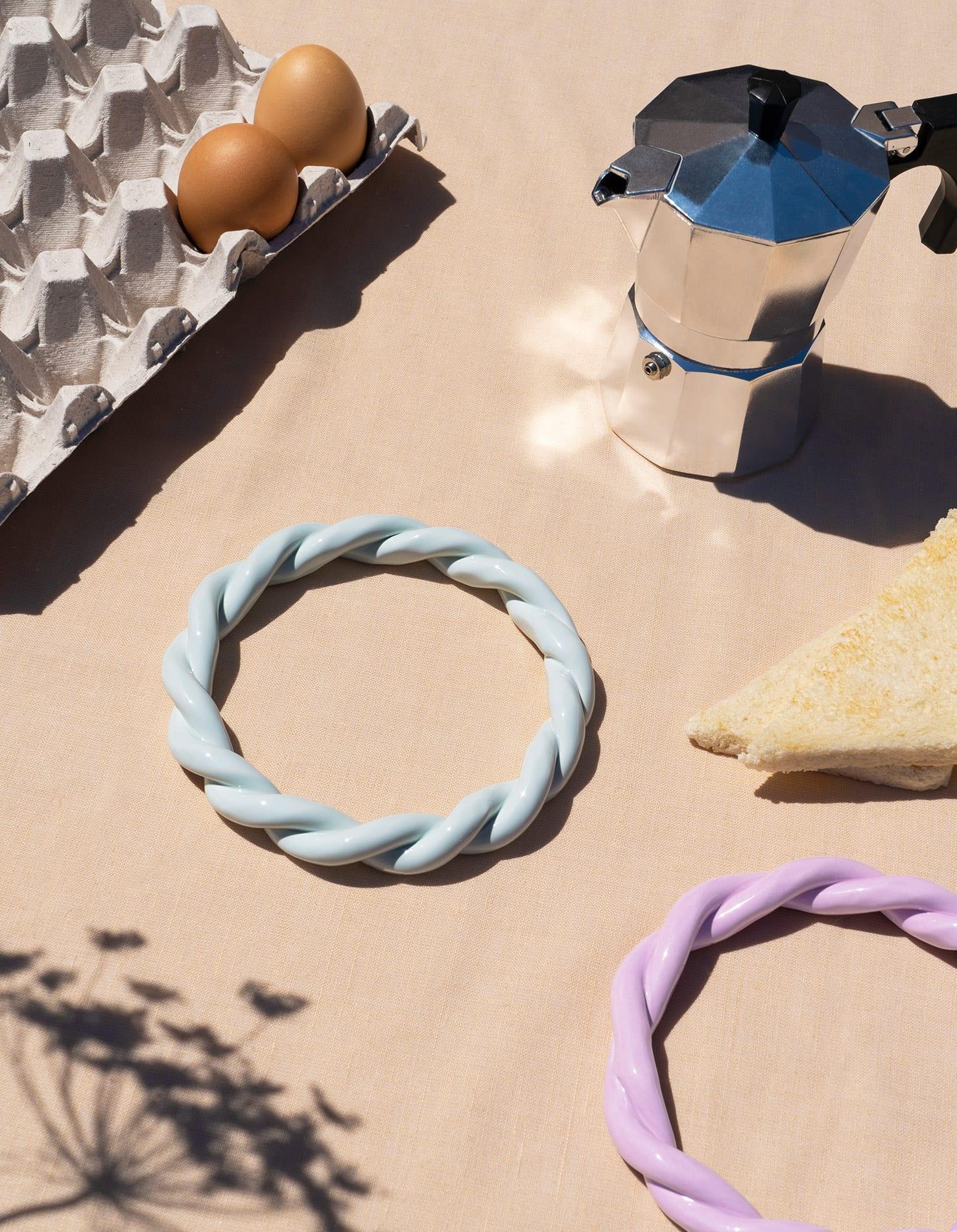 A moka pot, eggs, cheese wedge, and a light blue OCTAEVO Sicilia Ceramic Trivet on a beige tablecloth.