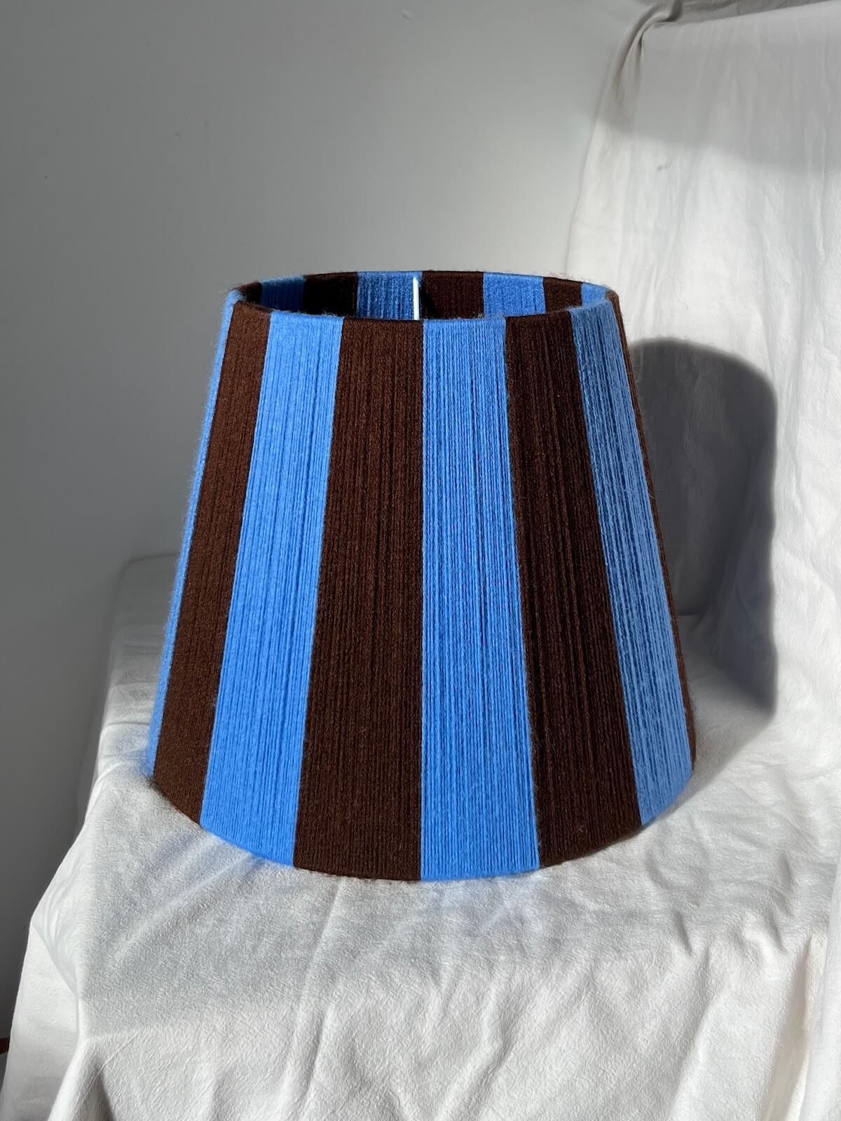 A Slice Shades handmade yarn striped lampshade with alternating vertical stripes of blue and dark brown, displayed on a white fabric backdrop.