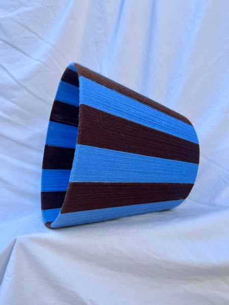 A Slice Shades handmade yarn striped lampshade with alternating vertical stripes of blue and dark brown, displayed on a white fabric backdrop.