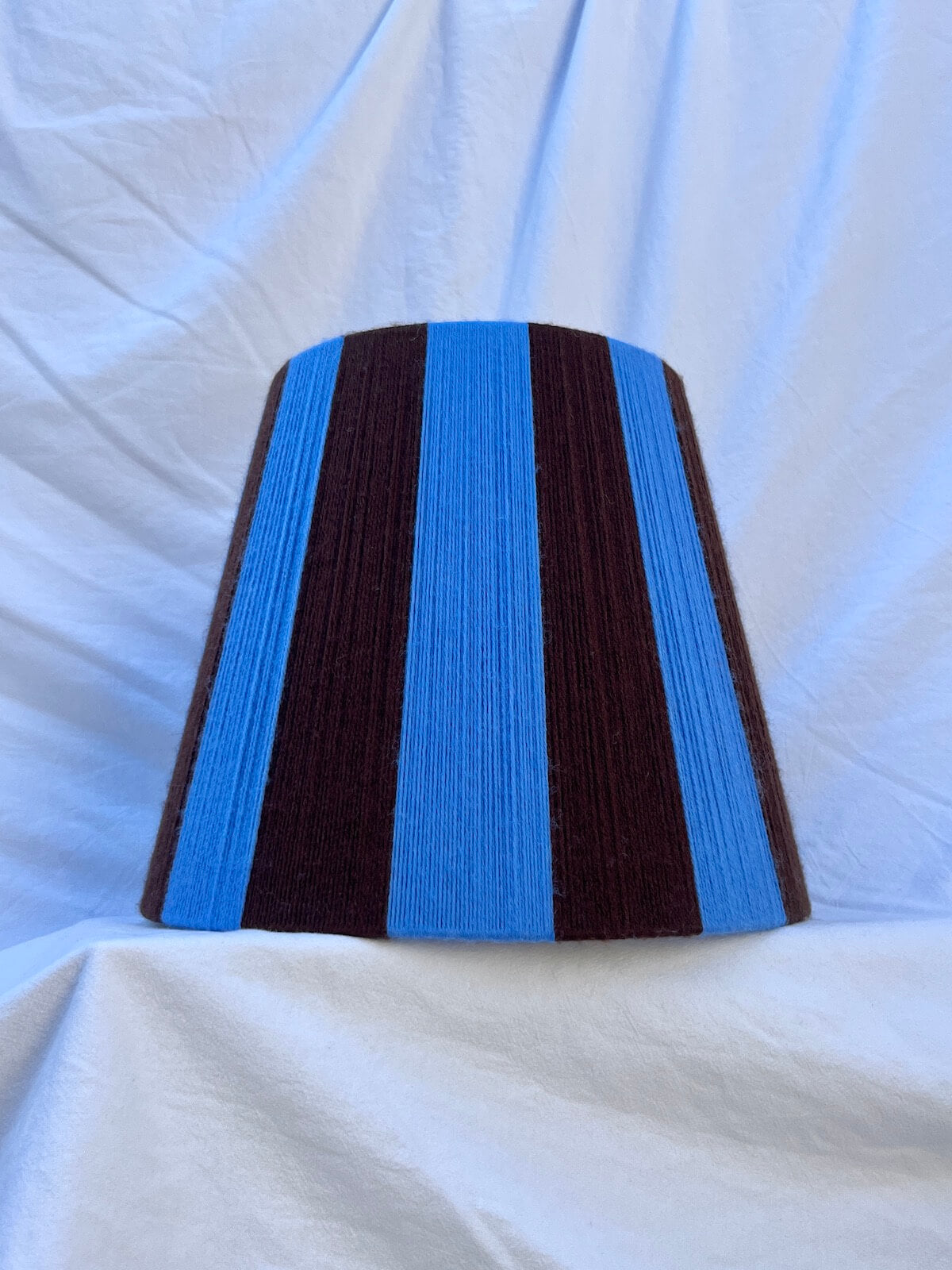 A Slice Shades handmade yarn striped lampshade with alternating vertical stripes of blue and dark brown, displayed on a white fabric backdrop.