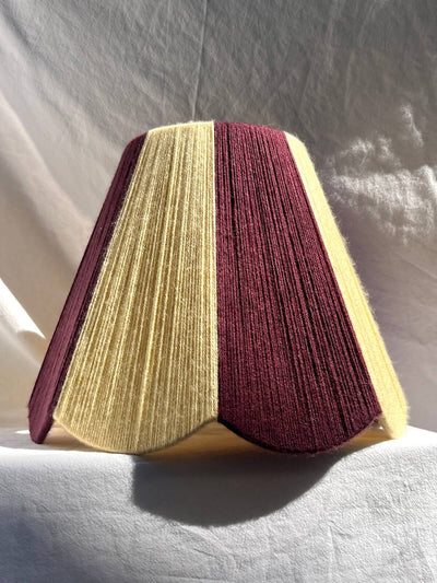 A Slice Shades lampshade with a handowoven yarn design of textured buttermilk yellow and deep burgundy, displayed on a draped white fabric background.