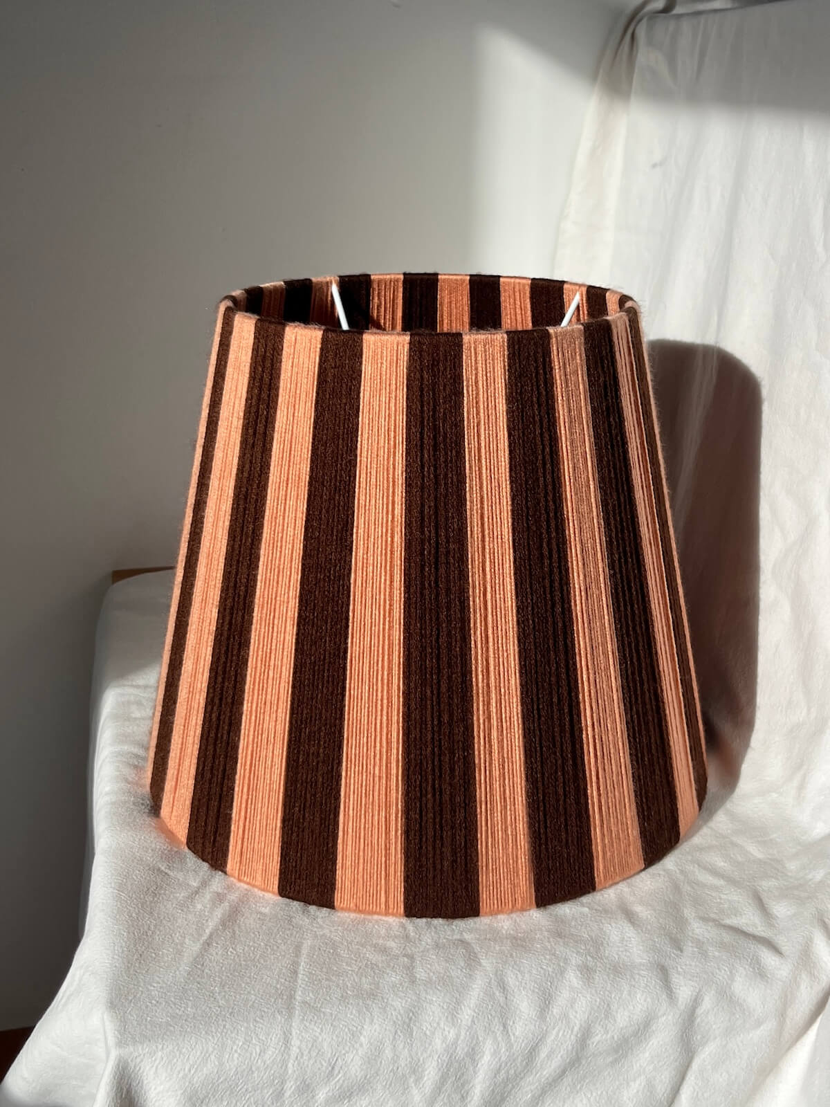 A Slice Shades handwoven striped yarn lampshade with alternating vertical stripes of dark brown and a warm peach-pink, displayed on a draped white fabric background in warm light.