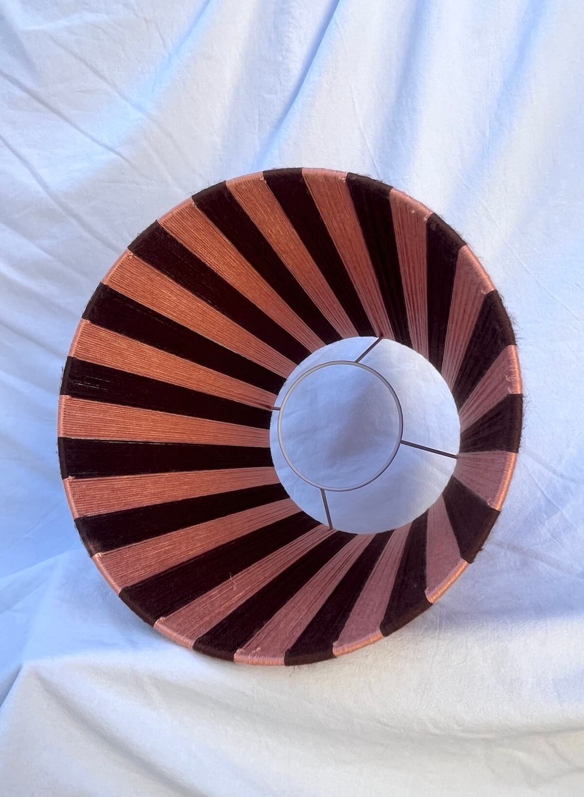 A Slice Shades handwoven striped yarn lampshade with alternating vertical stripes of dark brown and a warm peach-pink, displayed on a draped white fabric background inside view.
