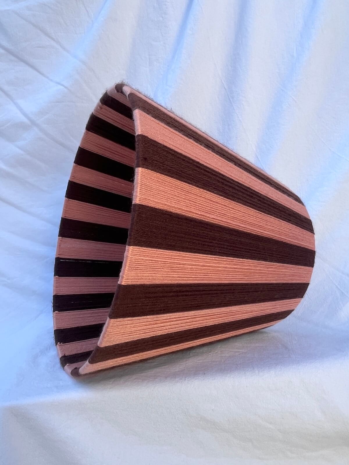 A Slice Shades handwoven striped yarn lampshade with alternating vertical stripes of dark brown and a warm peach-pink, displayed on a draped white fabric background on side.