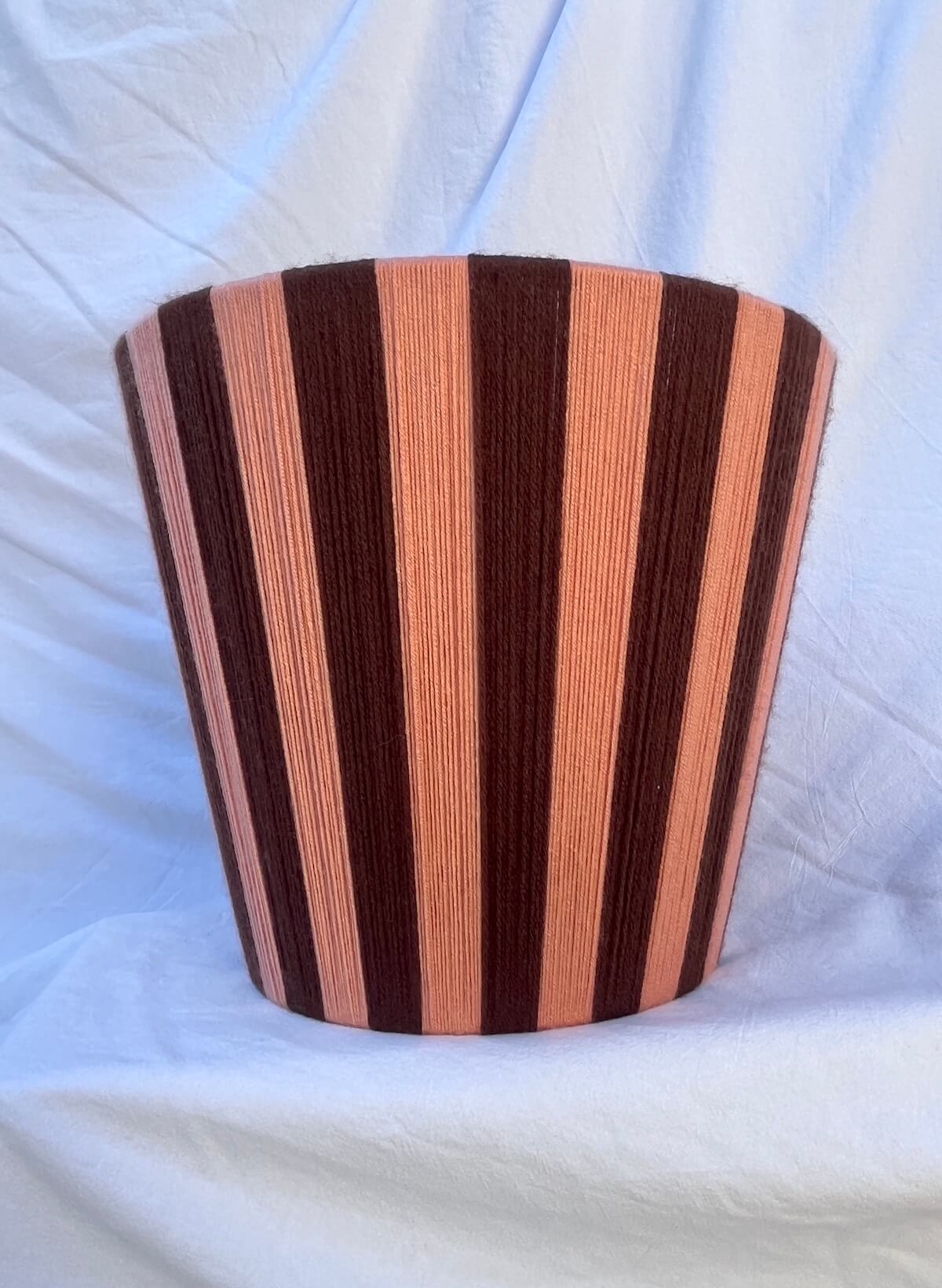 A Slice Shades handwoven striped yarn lampshade with alternating vertical stripes of dark brown and a warm peach-pink, displayed on a draped white fabric background.