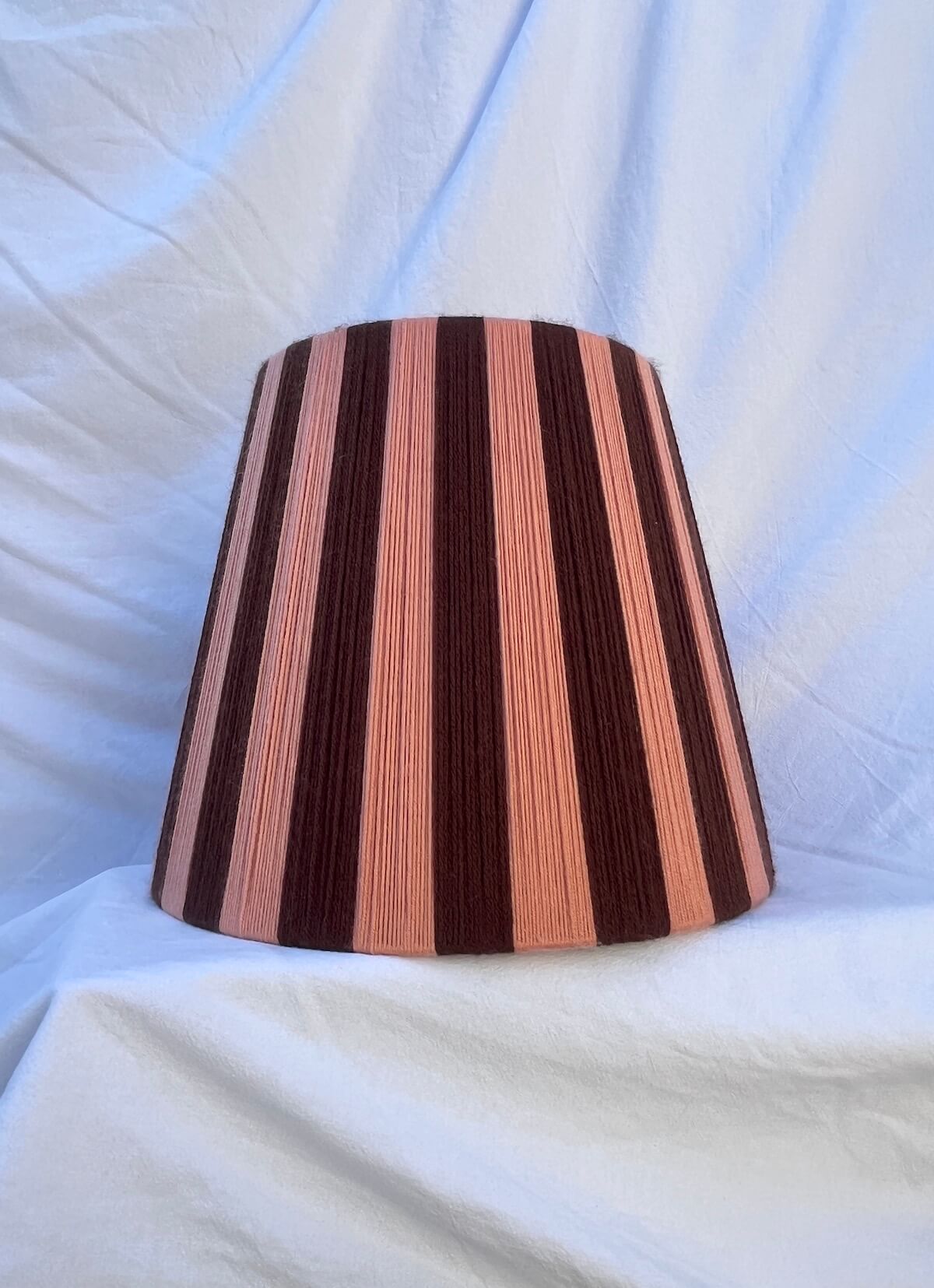 A Slice Shades handwoven striped yarn lampshade with alternating vertical stripes of dark brown and a warm peach-pink, displayed on a draped white fabric background.