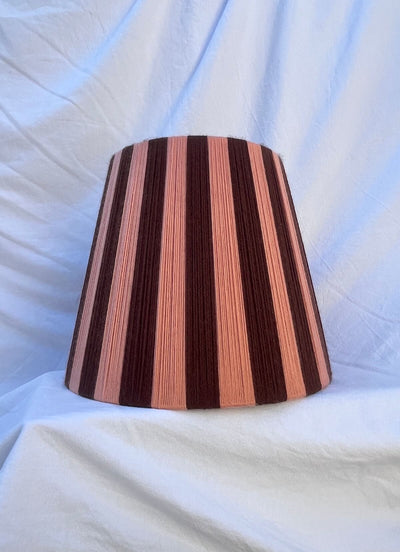 A Slice Shades handwoven striped yarn lampshade with alternating vertical stripes of dark brown and a warm peach-pink, displayed on a draped white fabric background.