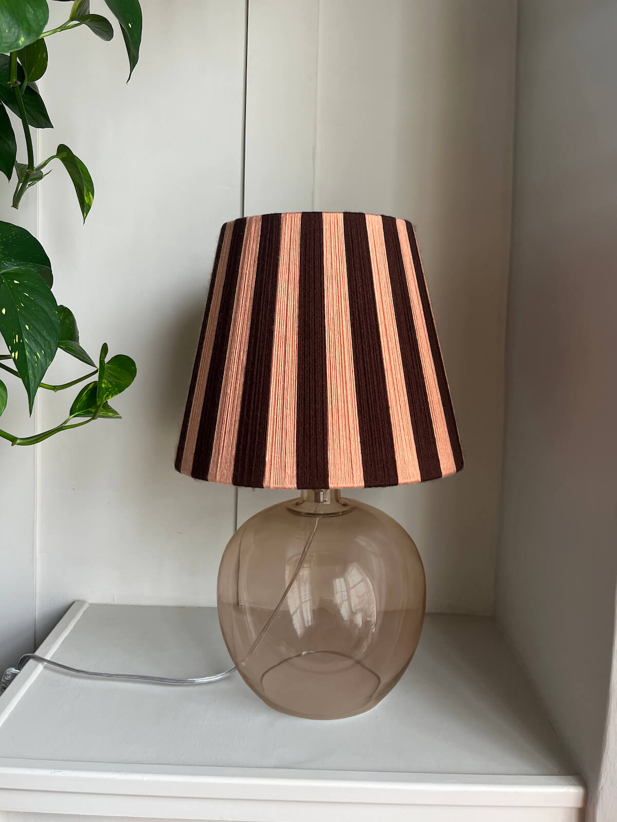 A Slice Shades handwoven striped yarn lampshade with alternating vertical stripes of dark brown and a warm peach-pink, mounted on a round, translucent smoky amber glass base and placed on a white shelf next to a green plant.