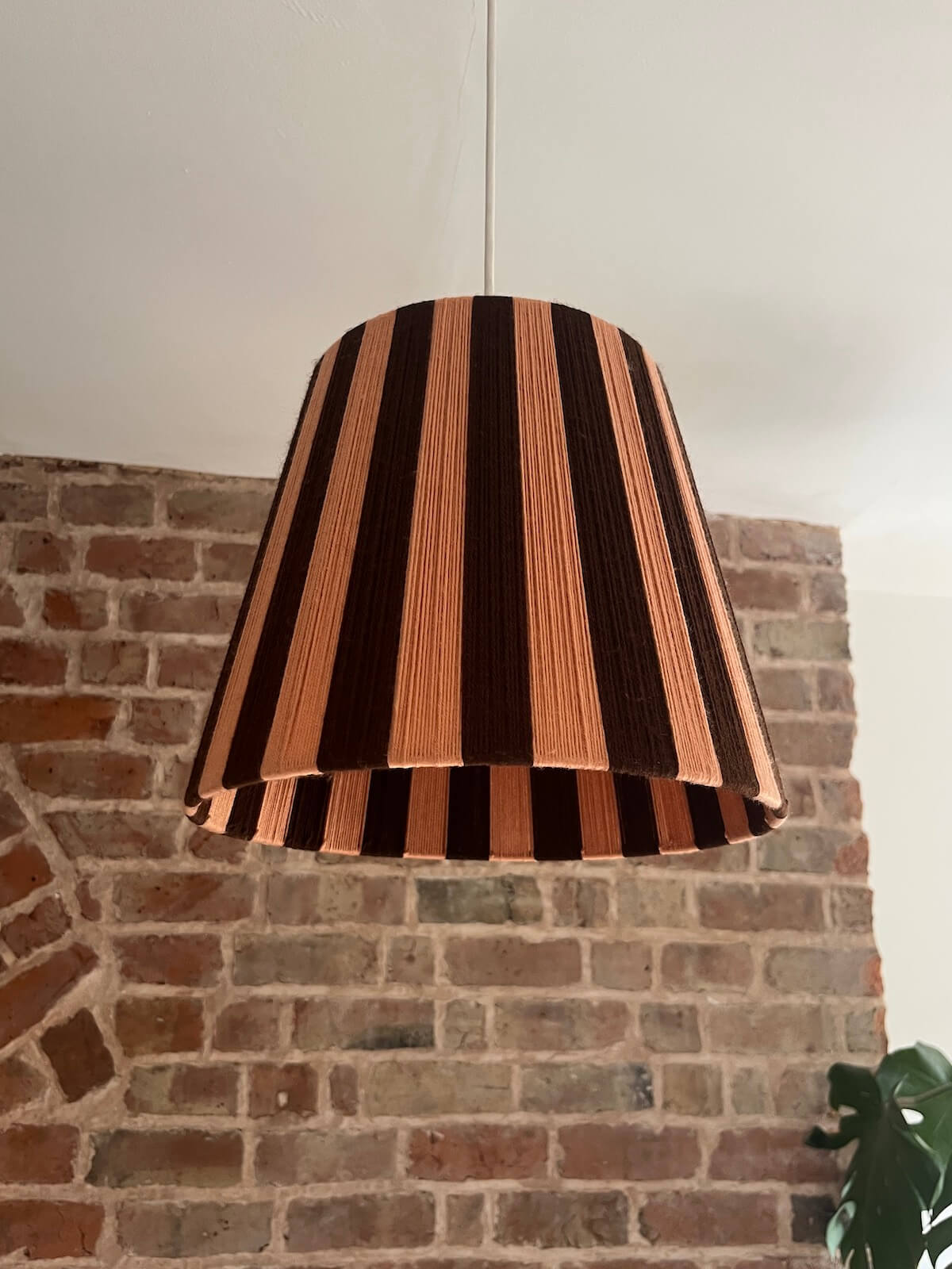 A Slice Shades handwoven striped yarn lampshade with alternating vertical stripes of dark brown and a warm peach-pink, hanging against a brick wall background.