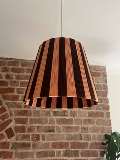A Slice Shades handwoven striped yarn lampshade with alternating vertical stripes of dark brown and a warm peach-pink, hanging against a brick wall background.