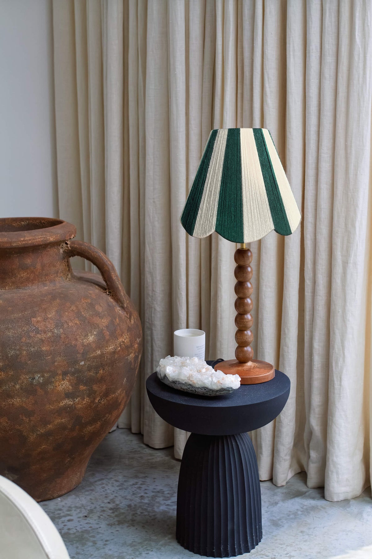 A Slice Shades handwoven yarn lampshade in green and cream striped table lamp with a wooden spindle base sits on a black pedestal side table, styled in a room with beige curtains, a large terracotta vase, and a white candle.