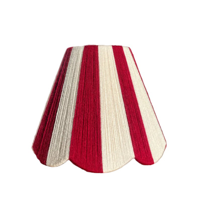 A Slice Shades yarn handwoven striped scalloped lampshade with alternating vertical stripes of deep red and off-white, displayed on a rustic wooden table with dramatic natural sunlight casting leaf shadows on the wall behind it.