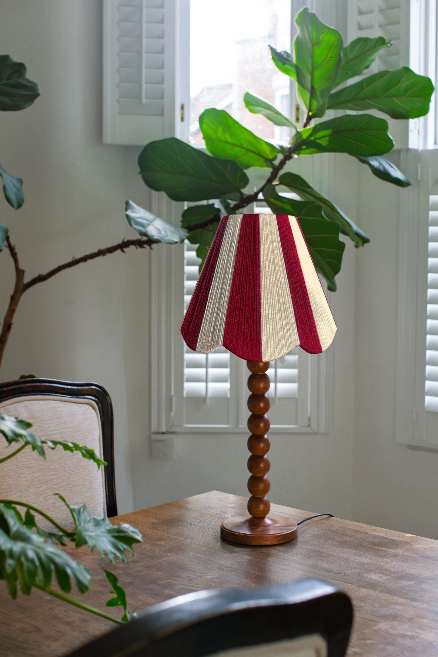 A Slice Shades yarn handwoven striped scalloped lampshade on a turned wood base, styled on a wooden dining table next to a fiddle-leaf fig plant in a bright room with white shutters.