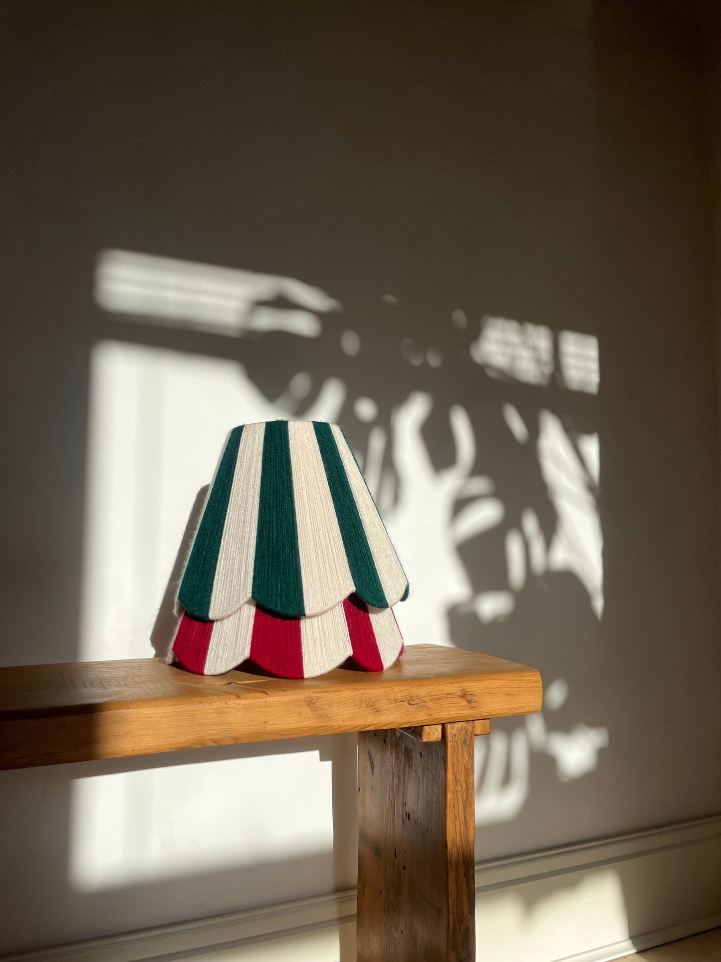 A Slice Shades yarn handwoven striped scalloped lampshade with stripes of deep red and off-white, layered over another shade with dark green and off-white stripes, displayed on a wooden table in warm natural light with shadows.