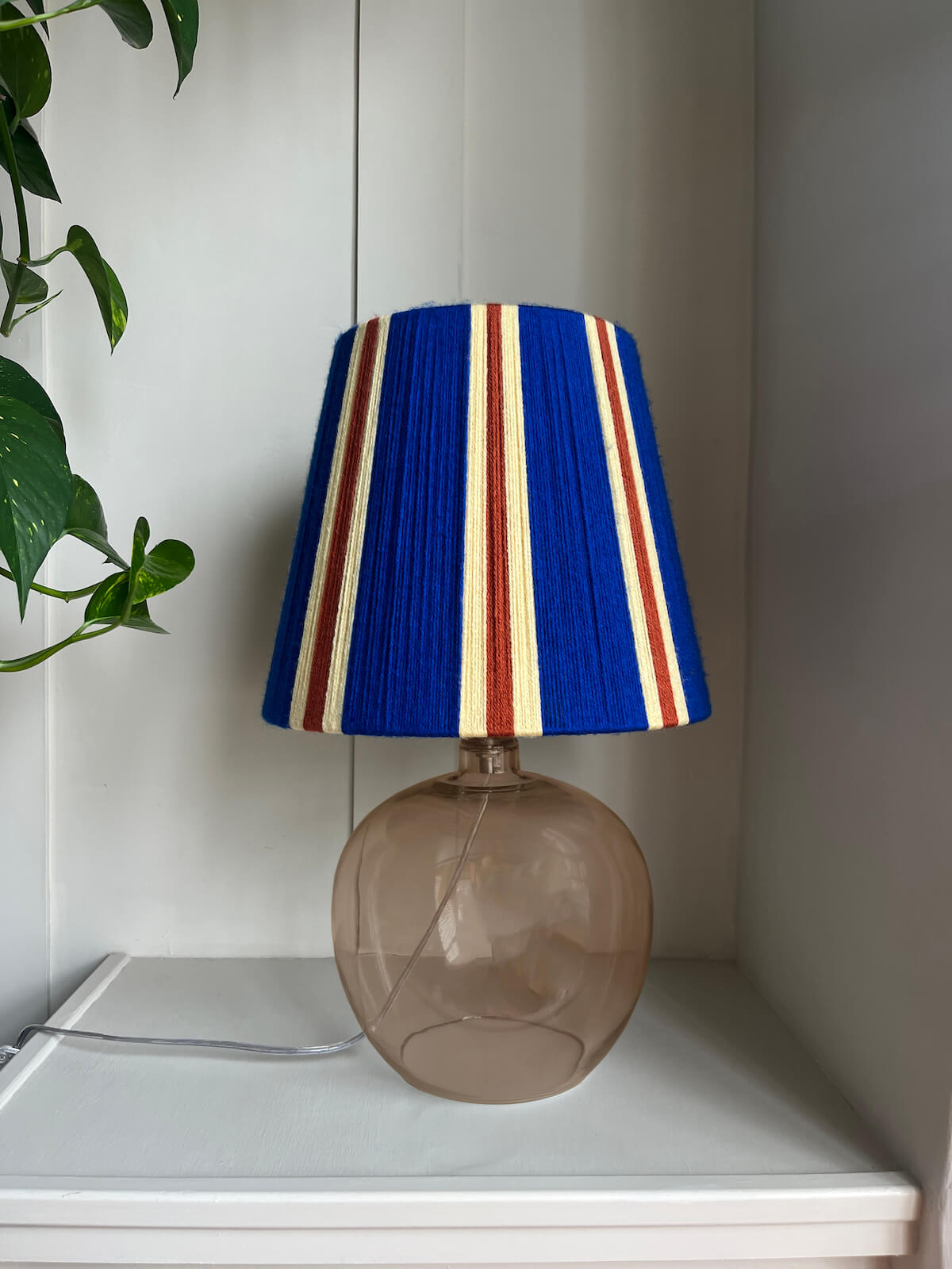 A Slice Shades handwoven striped yarn lampshade in cobalt blue, butter yellow and burnt orange stripes., paired with a round, translucent smoky brown glass base, displayed on a white shelf next to a green plant.