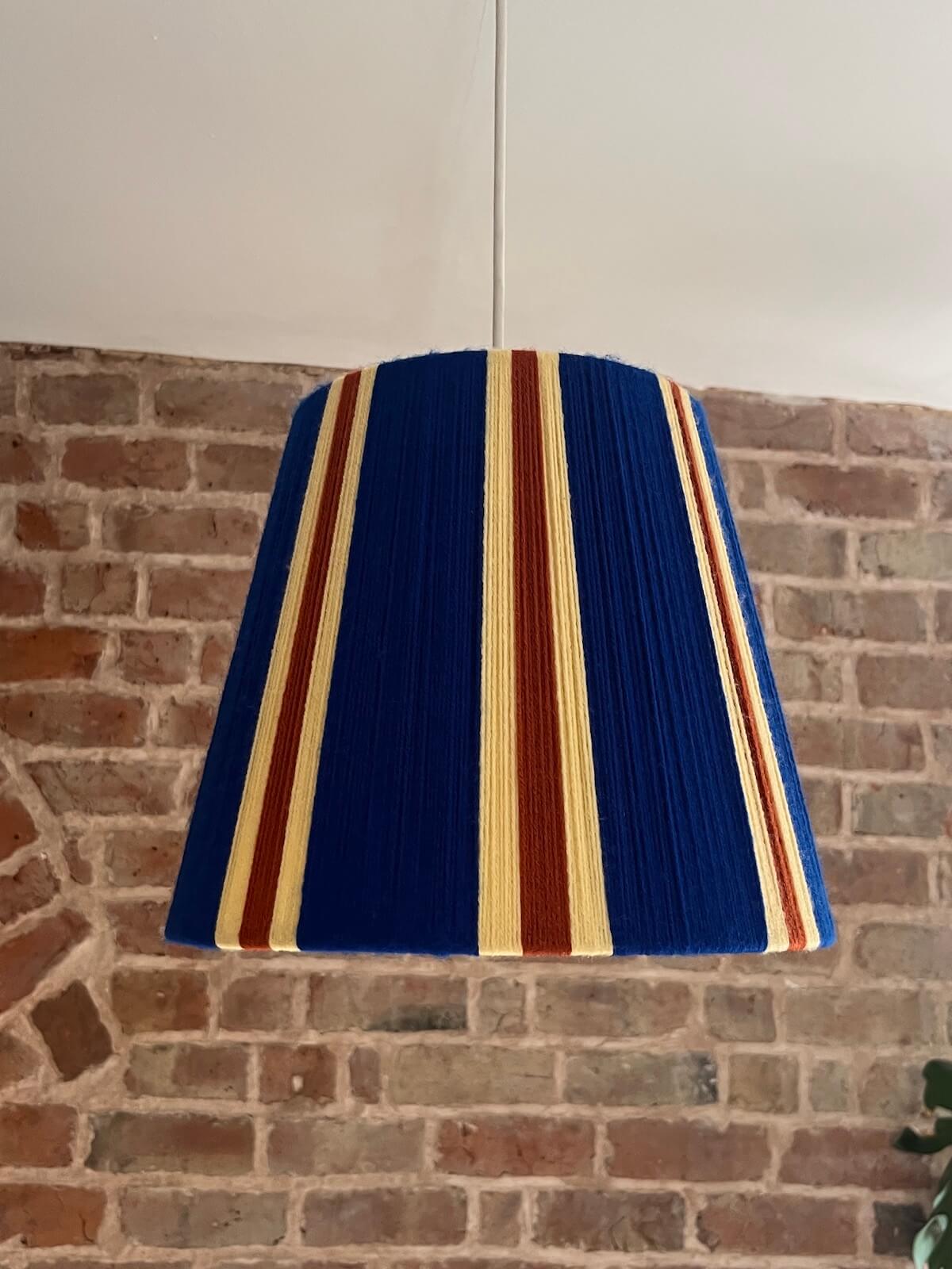 A Slice Shades handwoven striped yarn lampshade in cobalt blue, butter yellow and burnt orange stripes., hanging against a brick wall background.