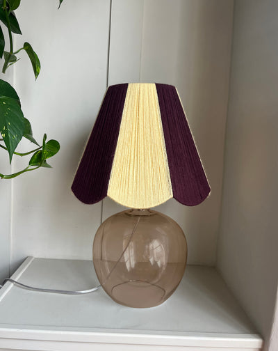 A table lamp with a Slice Shades lampshade with a handwoven yarn design of textured buttermilk yellow and deep burgundy, mounted on a round, transparent smoky-brown glass base, sitting on a white shelf next to a green plant.