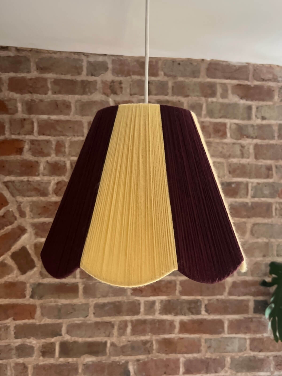 A Slice Shades lampshade with a handwoven yarn design of textured buttermilk yellow and deep burgundy, hanging against a rustic brick wall. 