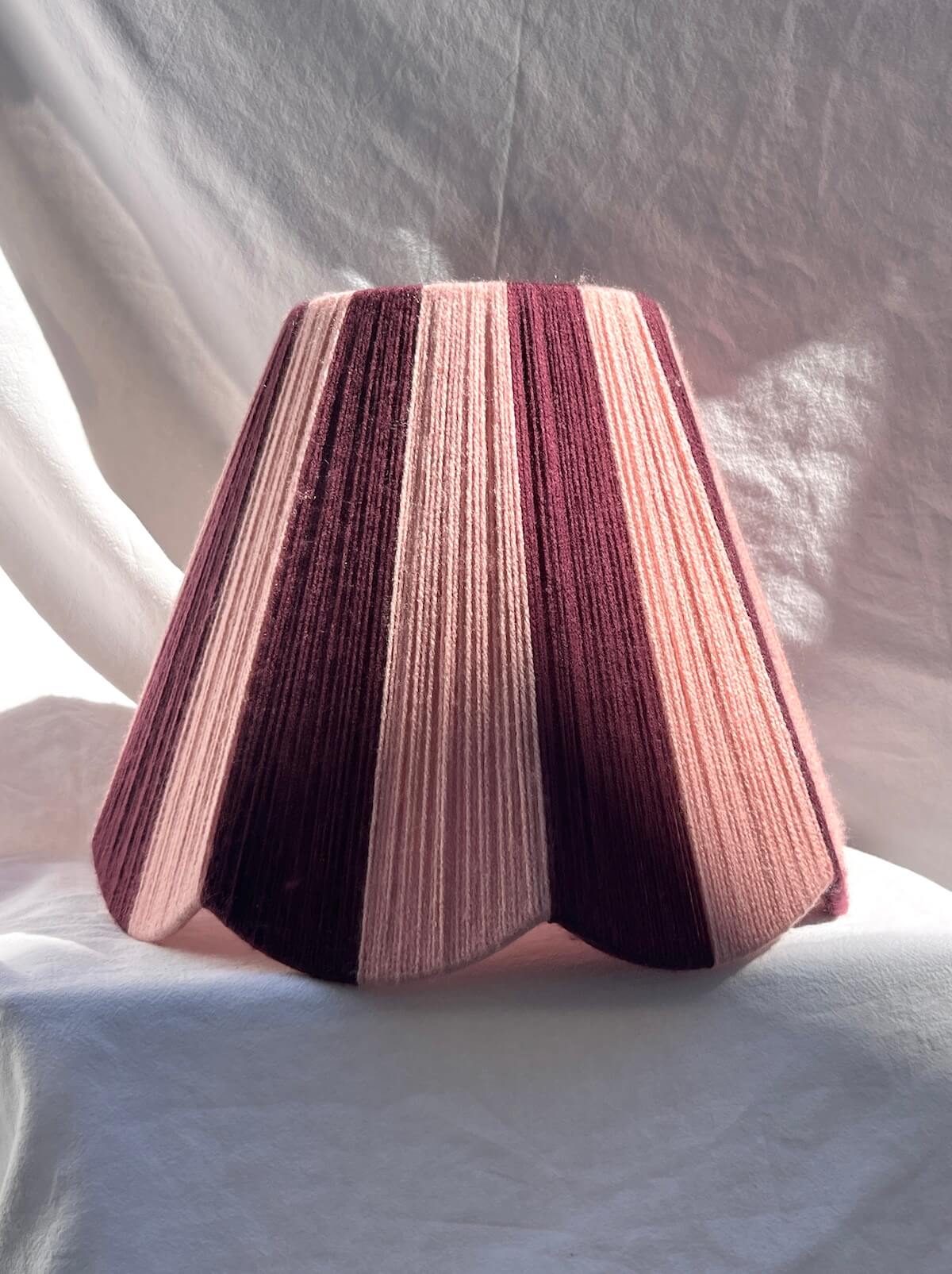 A Slice Shades handwoven yarn lampshade with vertical stripes in dusty pink  and deep burgundy, set against a draped white fabric background.