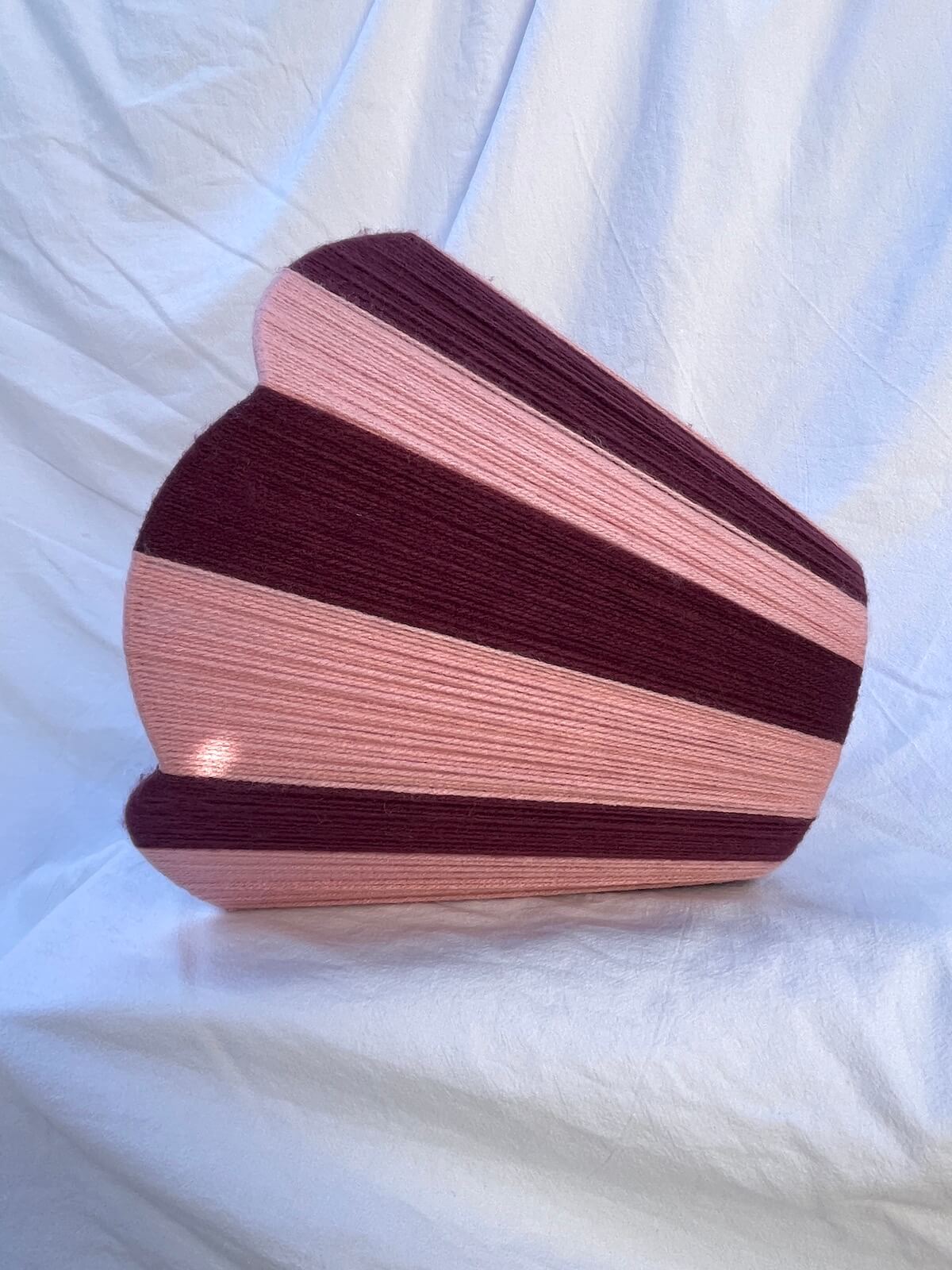A Slice Shades handwoven yarn lampshade with vertical stripes in dusty pink  and deep burgundy, set against a draped white fabric background.
