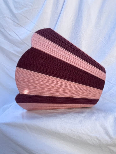 A Slice Shades handwoven yarn lampshade with vertical stripes in dusty pink  and deep burgundy, set against a draped white fabric background.