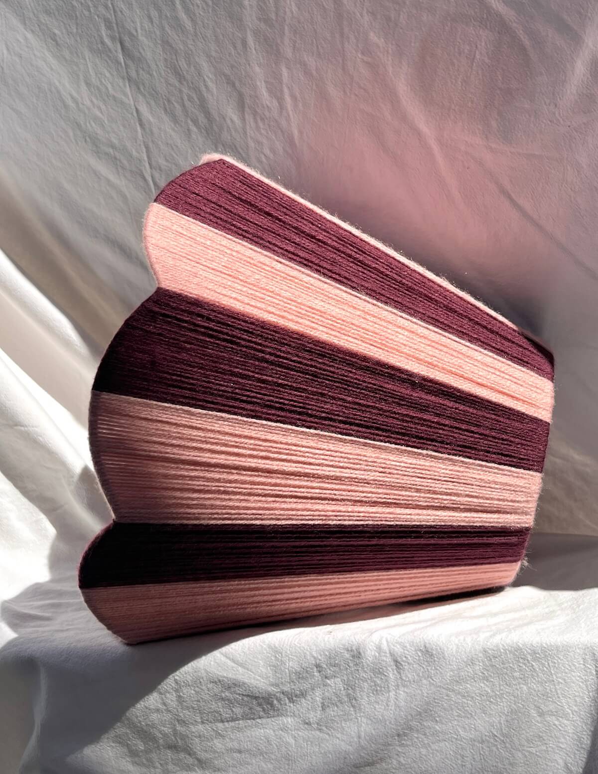 A Slice Shades handwoven yarn lampshade with vertical stripes in dusty pink  and deep burgundy, set against a draped white fabric background.