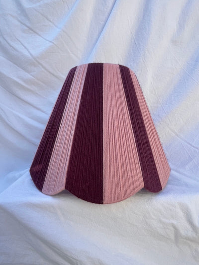 A Slice Shades handwoven yarn lampshade with vertical stripes in dusty pink  and deep burgundy, set against a draped white fabric background.