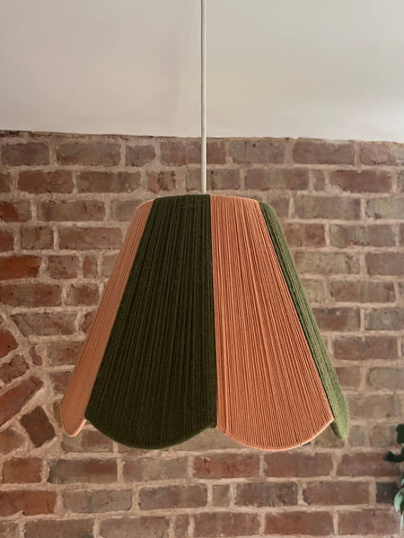 A slice shades handwoven striped scallop yarn lampshade in olive green and plaster pink, hanging against a brick wall background.