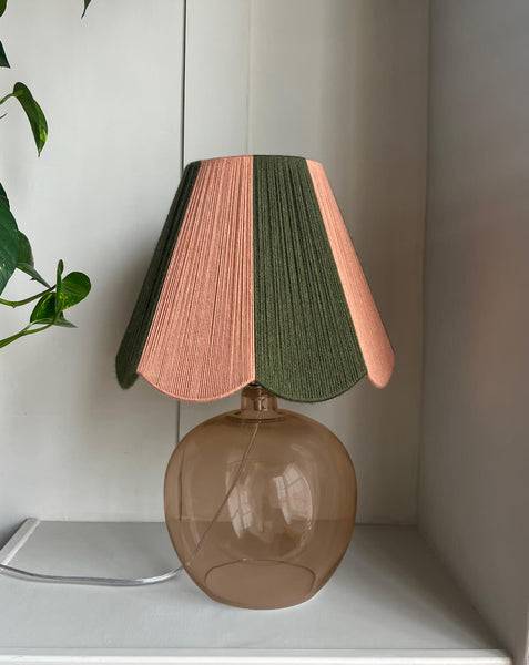 A table lamp with a slice shades handwoven striped scallop yarn lampshade in olive green and plaster pink, mounted on a rounded translucent smoky amber glass base, set on a white shelf next to a plant.