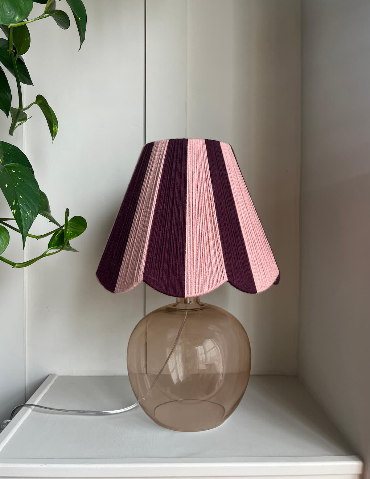 A Slice Shades handwoven yarn lampshade with vertical stripes in dusty pink  and deep burgundy table lamp with alternating pink and dark purple stripes on its shade, set on a white shelf next to a green plant.