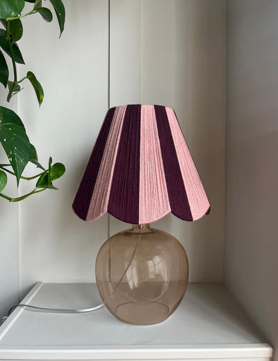 A Slice Shades handwoven yarn lampshade with vertical stripes in dusty pink  and deep burgundy table lamp with alternating pink and dark purple stripes on its shade, set on a white shelf next to a green plant.