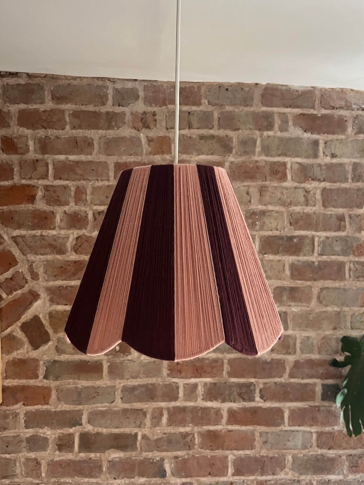 A Slice Shades handwoven yarn lampshade with vertical stripes in dusty pink  and deep burgundy, hanging against a rustic red-brown brick wall.