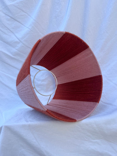 A Slice Shades handwoven yarn striped lampshade with vertical stripes in rust orange and light pink, photographed against a wrinkled white fabric backdrop.