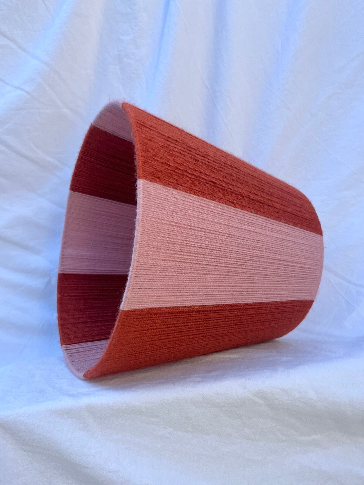A Slice Shades handwoven yarn striped lampshade with vertical stripes in rust orange and light pink, photographed against a wrinkled white fabric backdrop.