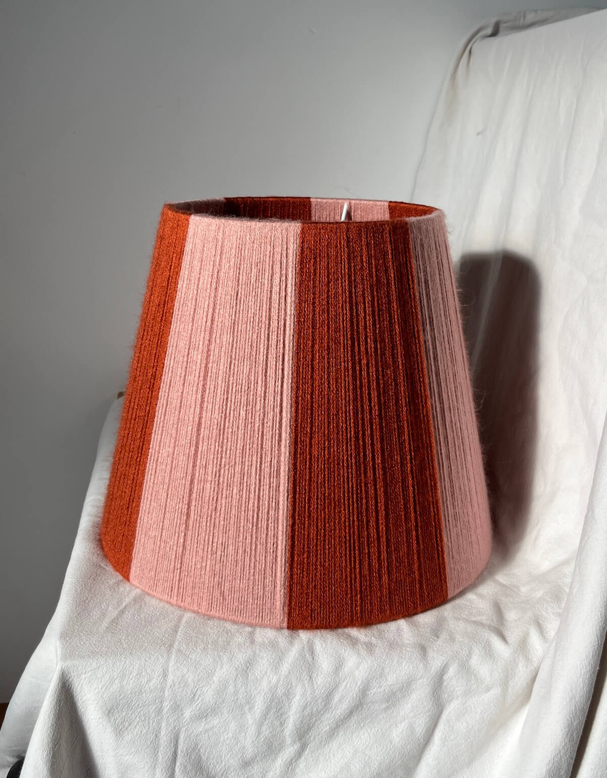 A Slice Shades handwoven yarn striped lampshade with vertical stripes in rust orange and light pink, photographed against a wrinkled white fabric backdrop.