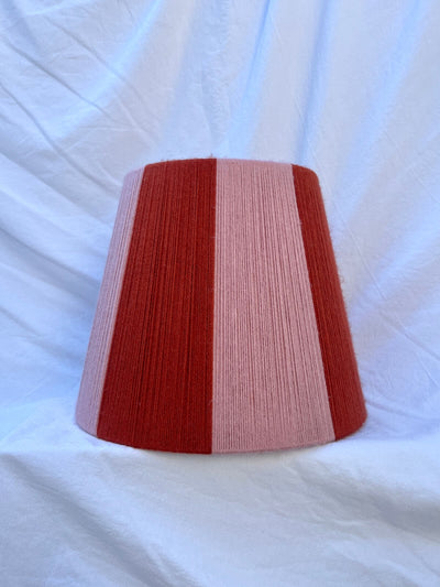 A Slice Shades handwoven yarn striped lampshade with vertical stripes in rust orange and light pink, photographed against a wrinkled white fabric backdrop.