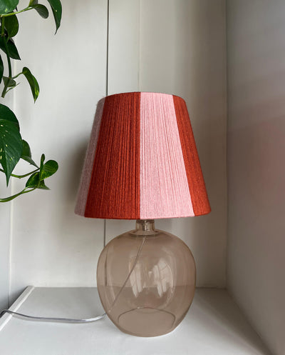 A table lamp with a Slice Shades handwoven yarn striped lampshade with vertical stripes in rust orange and light pink, mounted on a translucent smoky amber glass base, set against a white panelled wall with a green plant leaf visible on the left.