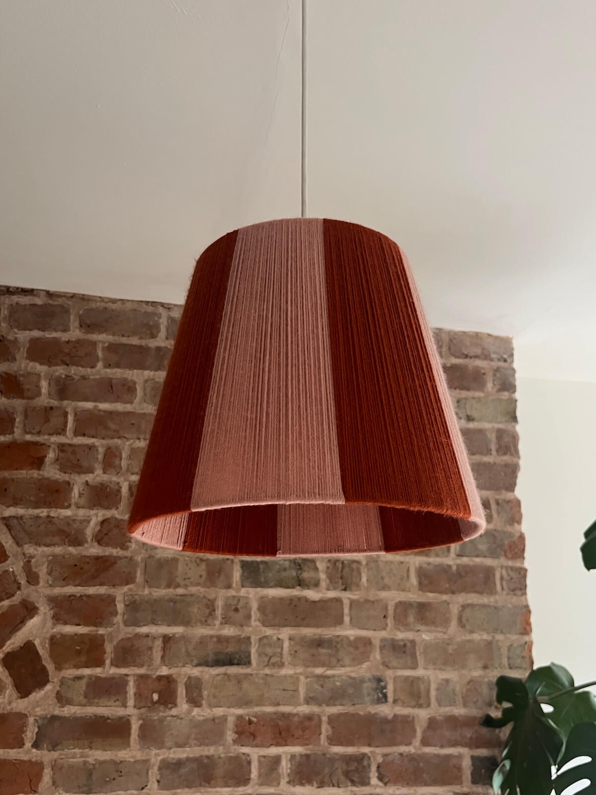 A Slice Shades handwoven yarn striped lampshade with vertical stripes in rust orange and light pink, hanging from a thin cord against a brick wall and white ceiling.