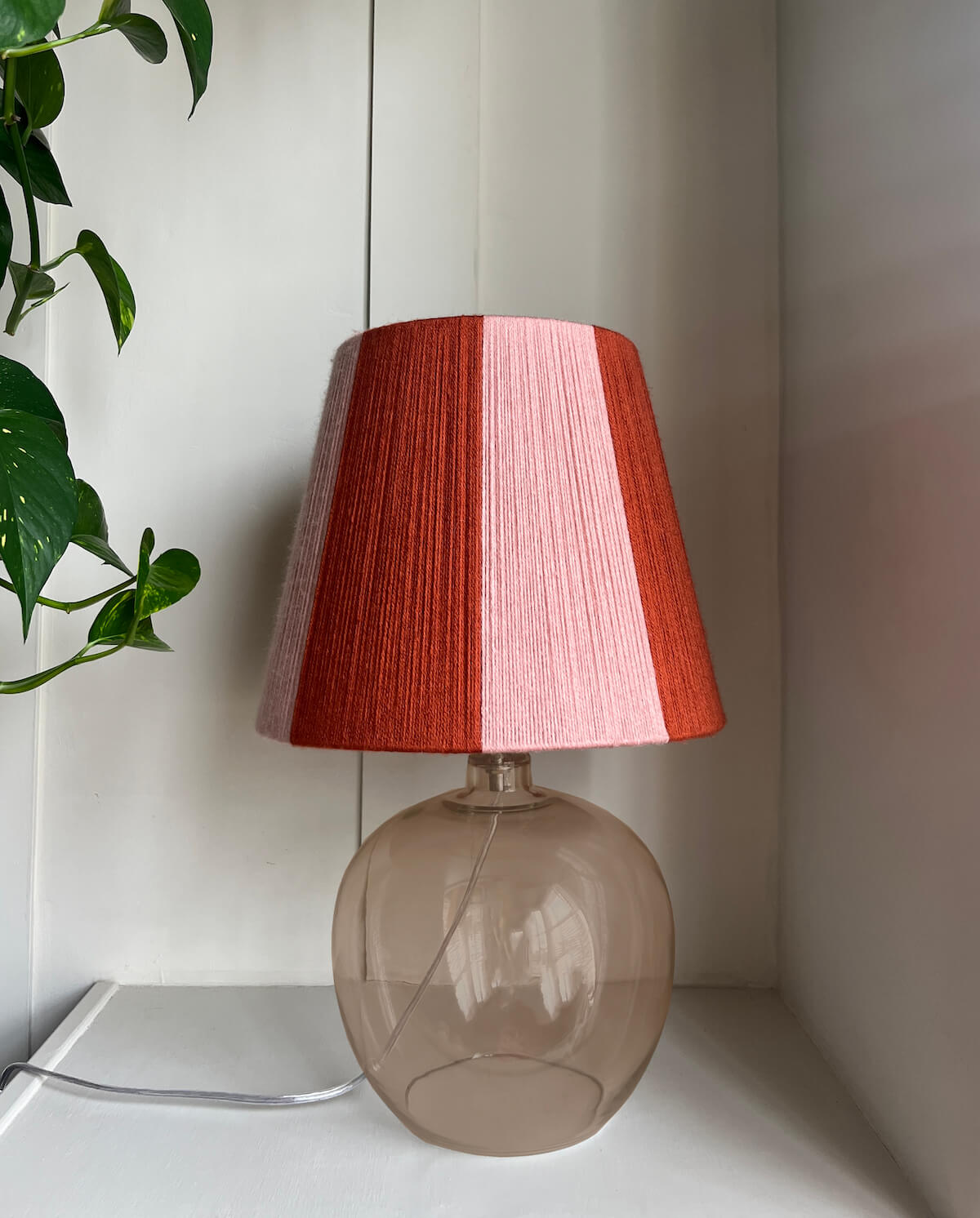 A table lamp with a Slice Shades handwoven yarn striped lampshade with vertical stripes in rust orange and light pink, mounted on a translucent smoky amber glass base, set against a white panelled wall with a green plant leaf visible on the left.