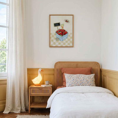 A styled bedroom interior featuring a single wooden bed with white bedding, a matching wooden headboard and footboard, a small bedside table with a lit lamp, cream curtains, and a framed artwork on the wall.