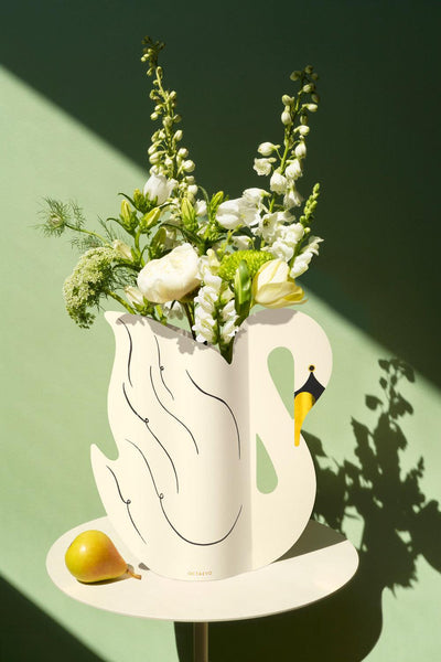 The Octaevo Swan Paper Vase with white flowers is displayed beside a yellow pear on a round table by a green wall.