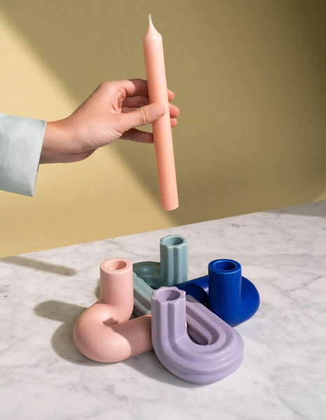 A hand holds a candle above OCTAEVO Templo Candle Holders, in pink, in blue, in purple and in light blue displayed on a marble surface.
