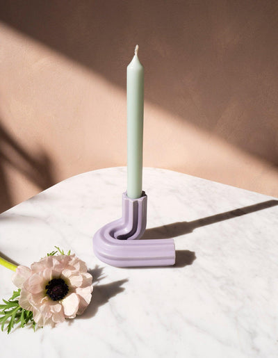 A pastel candle in OCTAEVO’s purple Templo Candle Holder, on marble, with a pale flower beside it.
