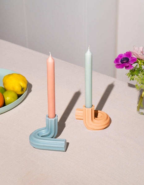 Two pastel candles in light blue OCTAEVO Templo Candle Holders on a table, with fruit and flowers in the background.